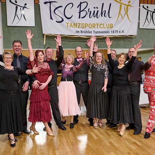 Gruppe von Tänzern in festlicher Kleidung mit erhobenen Händen vor einem Banner des TSC Brühl in einem Tanzsaal.