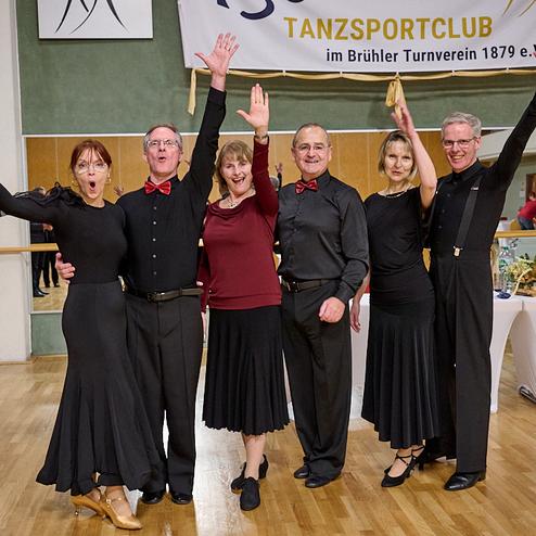 Sechs tanzende Personen in eleganter Kleidung jubeln in einem Tanzsaal mit Spiegelwand und festlicher Dekoration.