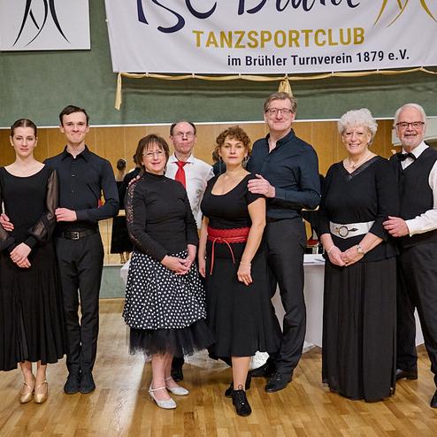 Gruppe von Tanzsportlern in formeller Kleidung steht vor einem Spiegel, im Hintergrund ein Tisch mit Computern.
