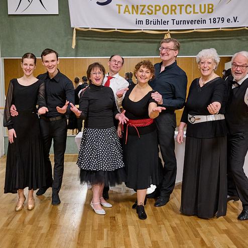 Gruppe von Tänzern in eleganter Kleidung posiert lächelnd in einem Tanzsaal mit spiegelnden Wänden.