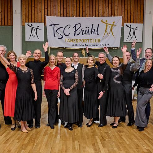 Gruppenselfie von Tänzern des TSC Brühl in festlicher Kleidung, lächelnd vor einem Vereinsbanner.