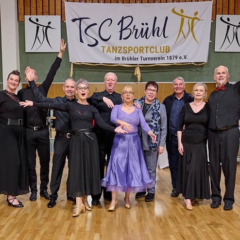 Gruppe von Tänzern in eleganter Kleidung posiert vor einem Banner des TSC Brühl im Tanzsportclub.