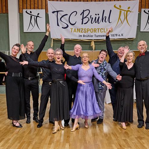 Gruppenszene mit Tänzern in festlicher Kleidung, die vor einem Banner des TSC Brühl posieren und Freude zeigen.