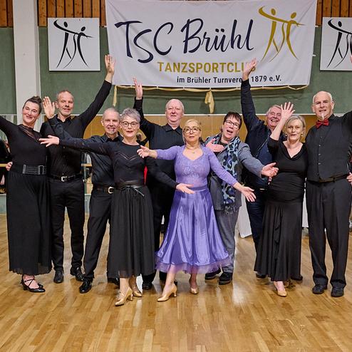 Gruppe von Tänzern in eleganter Abendkleidung, die freudig in einem Tanzsaal posieren, vor einem Banner des Tanzsportclubs.