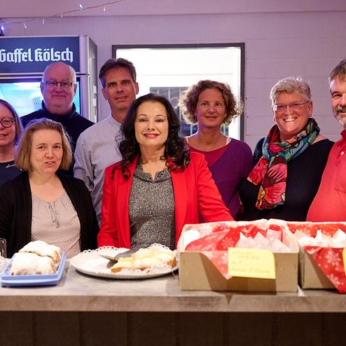 Gruppenshot von acht Personen lächelnd hinter einem Tisch mit Gebäck und Snacks in einem festlichen Raum.