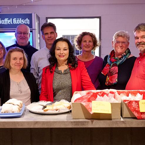 Gruppe von acht Personen lächelt vor einem Stehtisch mit verschiedenen Speisen und Getränken.