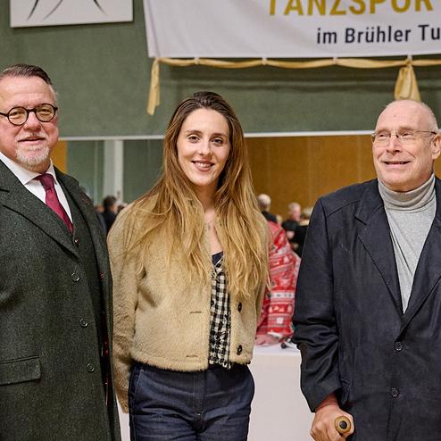 Drei Personen posieren lächelnd vor einem Banner im Hintergrund, das auf einen Tanzsportverein hinweist.