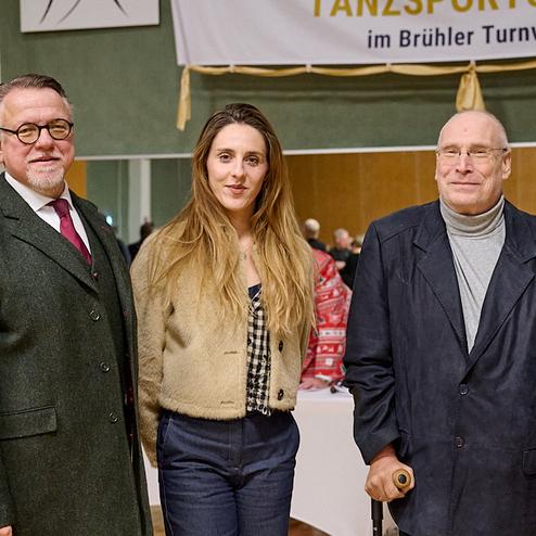 Drei Personen stehen lächelnd in einem Raum mit festlicher Dekoration im Hintergrund.