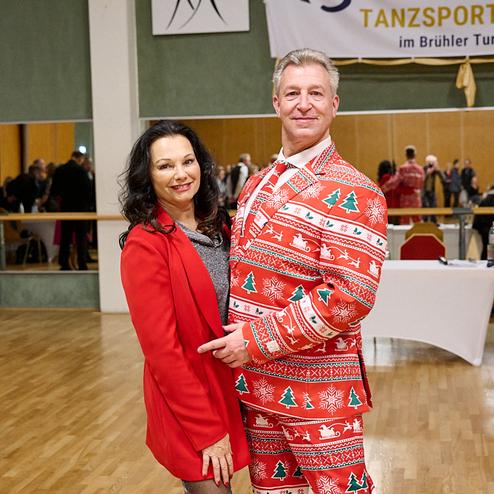 Paar lächelt in festlicher Kleidung, der Mann trägt einen bunten Anzug mit Weihnachtsmotiven, im Hintergrund Tanzveranstaltung.