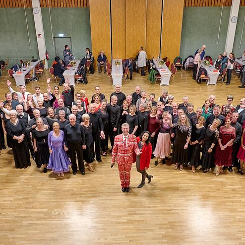 Gruppensfoto von Tänzern in eleganter Kleidung, die auf einer Tanzfläche zusammenstehen. Tische im Hintergrund.