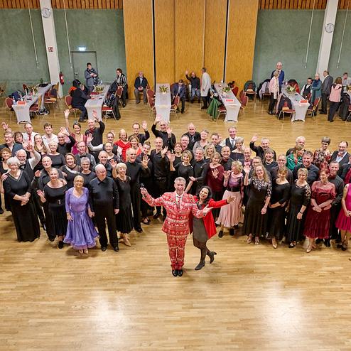 Gruppengestellte Tänzer in festlicher Kleidung lächeln und winken in einer großen Tanzfläche mit Tischdekoration.
