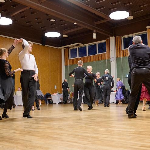 Tanzende Paare in eleganter Kleidung auf einer Holzfläche, umgeben von Tischen in einem festlichen Saal.