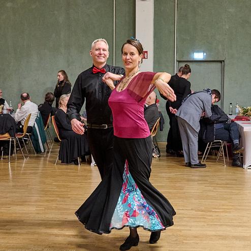Tanzendes Paar in eleganter Kleidung, umgeben von Gästen in einem festlichen Saal mit Holzwänden.