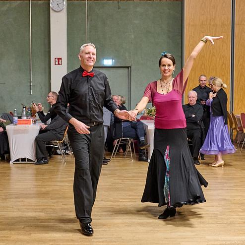 Tanzendes Paar in eleganter Kleidung auf der Tanzfläche, umgeben von Gästen in einem festlichen Saal.