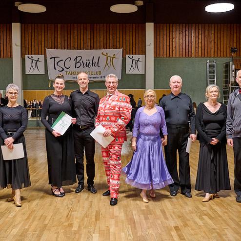 Gruppe von Tänzern in eleganter Kleidung steht auf einer Tanzfläche, mit Urkunden in der Hand und einem Banner im Hintergrund.