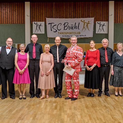 Gruppe von zwölf Tänzern in festlicher Kleidung, posierend vor einem Banner des TSC Brühl in einer Tanzhalle.