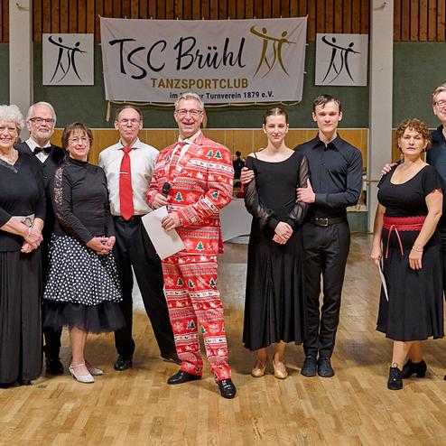 Gruppe von Tänzern in festlicher Kleidung steht vor einem Tanzsaal mit TSC Brühl Banner im Hintergrund.