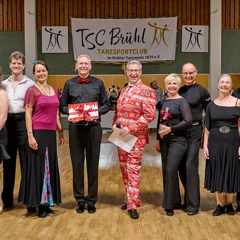 Gruppe von Tänzern in festlicher Kleidung posiert mit Pokalen vor einem Banner des TSC Brühl in einer Tanzhalle.