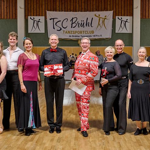 Gruppe von Tänzern in festlicher Kleidung posiert lächelnd in einem Tanzsaal mit einem Banner des TSC Brühl im Hintergrund.
