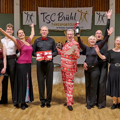 Gruppe von Tänzern in festlicher Kleidung, die strahlend auf einer Tanzfläche zusammen feiern. Banner im Hintergrund.