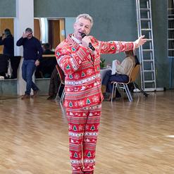 Mann in weihnachtlichem Anzug h&auml;lt Mikrofon und singt auf einer Tanzfl&auml;che, im Hintergrund G&auml;ste und St&uuml;hle.