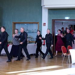 Tanzende Paare in eleganter Kleidung betreten einen Saal, Zuschauer sitzen an Tischen im Hintergrund.