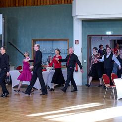 Tanzende Paare in eleganter Kleidung bewegen sich in einem festlichen Saal mit Tischen im Hintergrund.