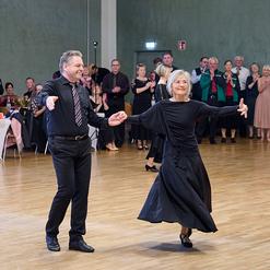 Tanzendes Paar in eleganter Kleidung, umgeben von applaudierendem Publikum in einem festlichen Saal.