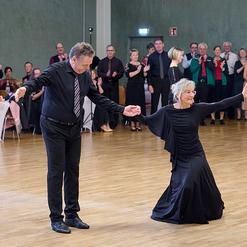 Tanzpaar in festlicher Kleidung pr&auml;sentiert eine Darbietung vor applaudierender Menge in einem Veranstaltungsraum.