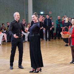 Tanzpaar in festlicher Kleidung, eine Frau winkt, w&auml;hrend eine andere mit einem Korb im Hintergrund l&auml;chelt. Zuschauer beobachten.