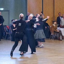 Tanzpaar in eleganter Kleidung auf einer Veranstaltung, umgeben von Zuschauern, die applaudieren und zuschauen.