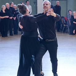 Tanzpaar in eleganter Kleidung f&uuml;hrt einen Tanz auf einer Halle mit Zuschauern im Hintergrund aus.