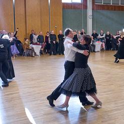 Tanzpaar im Halbprofil auf einer Tanzfl&auml;che, umgeben von Zuschauern in festlicher Kleidung.