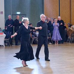 Paar tanzt in festlicher Kleidung auf einer Tanzfl&auml;che, umgeben von applaudierenden Zuschauern in einem Veranstaltungssaal.