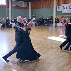 Tanzpaare in eleganter Kleidung tanzen auf einer hellen Holzfl&auml;che w&auml;hrend einer Tanzveranstaltung im Hintergrund.