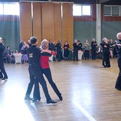 Tanzpaar in eleganter Kleidung tanzt auf einer Holzfl&auml;che, umgeben von Zuschauern in einem festlichen Saal.