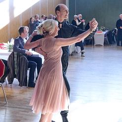 Tanzendes Paar auf einer Veranstaltung, umgeben von Publikum, in einem hellen Saal mit Holzvert&auml;felung.
