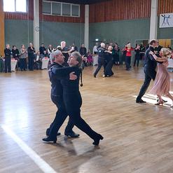 Tanzpaare in eleganter Kleidung beim Tanzen in einem festlichen Saal mit Publikum im Hintergrund.