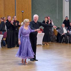 Tanzpaar in eleganter Kleidung bei einem Tanzabend, umgeben von applaudierendem Publikum in einem festlichen Saal.