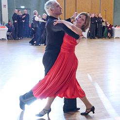 Tanzpaar in eleganter Kleidung f&uuml;hrt einen Tanz in einem festlichen Saal mit anderen T&auml;nzern im Hintergrund aus.