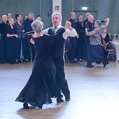 Tanzendes Paar in eleganter Kleidung auf einer Tanzfl&auml;che, umgeben von Zuschauern in festlicher Kleidung.