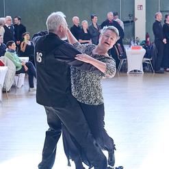 Tanzendes Paar in eleganter Kleidung auf einer Tanzveranstaltung, mit Zuschauern im Hintergrund.