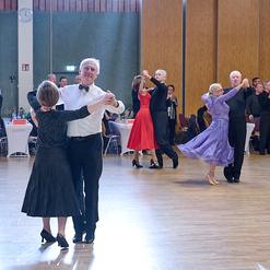 Tanzpaare in eleganter Kleidung tanzen auf einer Tanzfl&auml;che, G&auml;ste beobachten und applaudieren im Hintergrund.