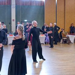 Tanzpaare in eleganter Kleidung auf einer Tanzfl&auml;che, im Hintergrund Zuschauer an Tischen.