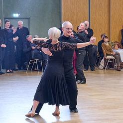 Tanzpaar in eleganter Kleidung tanzt in einem Saal, Zuschauer im Hintergrund beobachten aufmerksam.