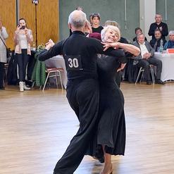 Tanzendes Paar in eleganter Kleidung auf einer Tanzfl&auml;che, umgeben von Zuschauern in einem Festsaal.