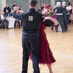 Tanzpaar in festlicher Kleidung f&uuml;hrt einen modernen Tanz in einem Saal mit Zuschauern im Hintergrund aus.
