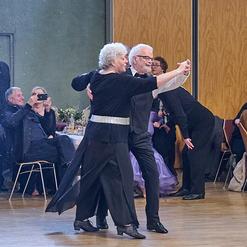 Tanzendes Paar in eleganter Kleidung auf einer Tanzfl&auml;che, mit anderen G&auml;sten im Hintergrund, die zuschauen.
