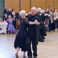 Tanzendes Paar im Schwarzen, umgeben von Zuschauern in einem festlichen Saal. Holzfu&szlig;boden und gro&szlig;e Fenster im Hintergrund.