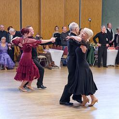 Tanzende Paare in eleganter Kleidung auf einer Holzfl&auml;che, umgeben von applaudierenden Zuschauern.